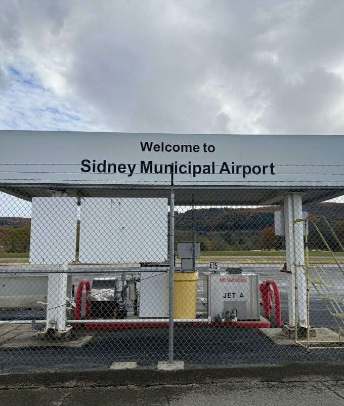 The fuel island and welcome sign at Sidney Municipal Airport, seen through the perimeter fence